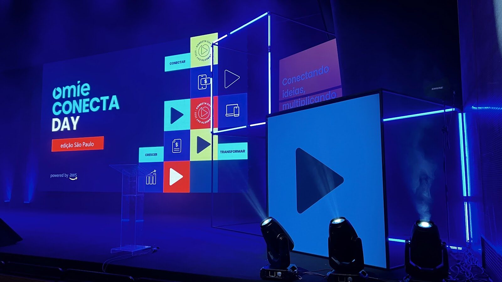Palco com cubo de neon, telão LED e moving heads em conferência corporativa