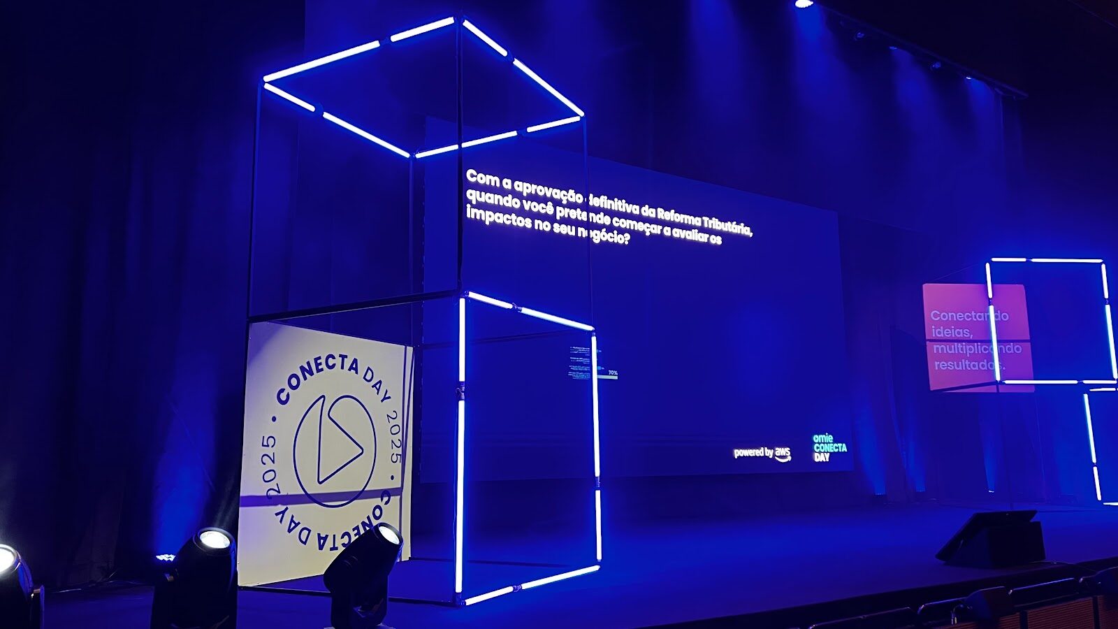 Palco de evento corporativo com painel LED azul, cubo de neon e cenografia integrada em ambiente escuro