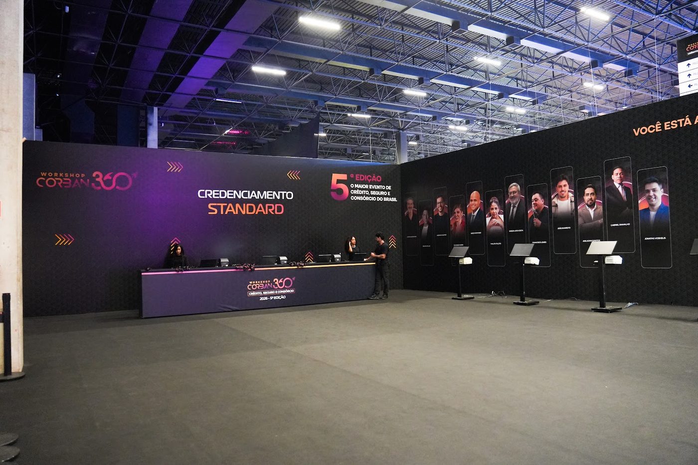 Balcão de credenciamento com painel de palestrantes iluminado em evento corporativo