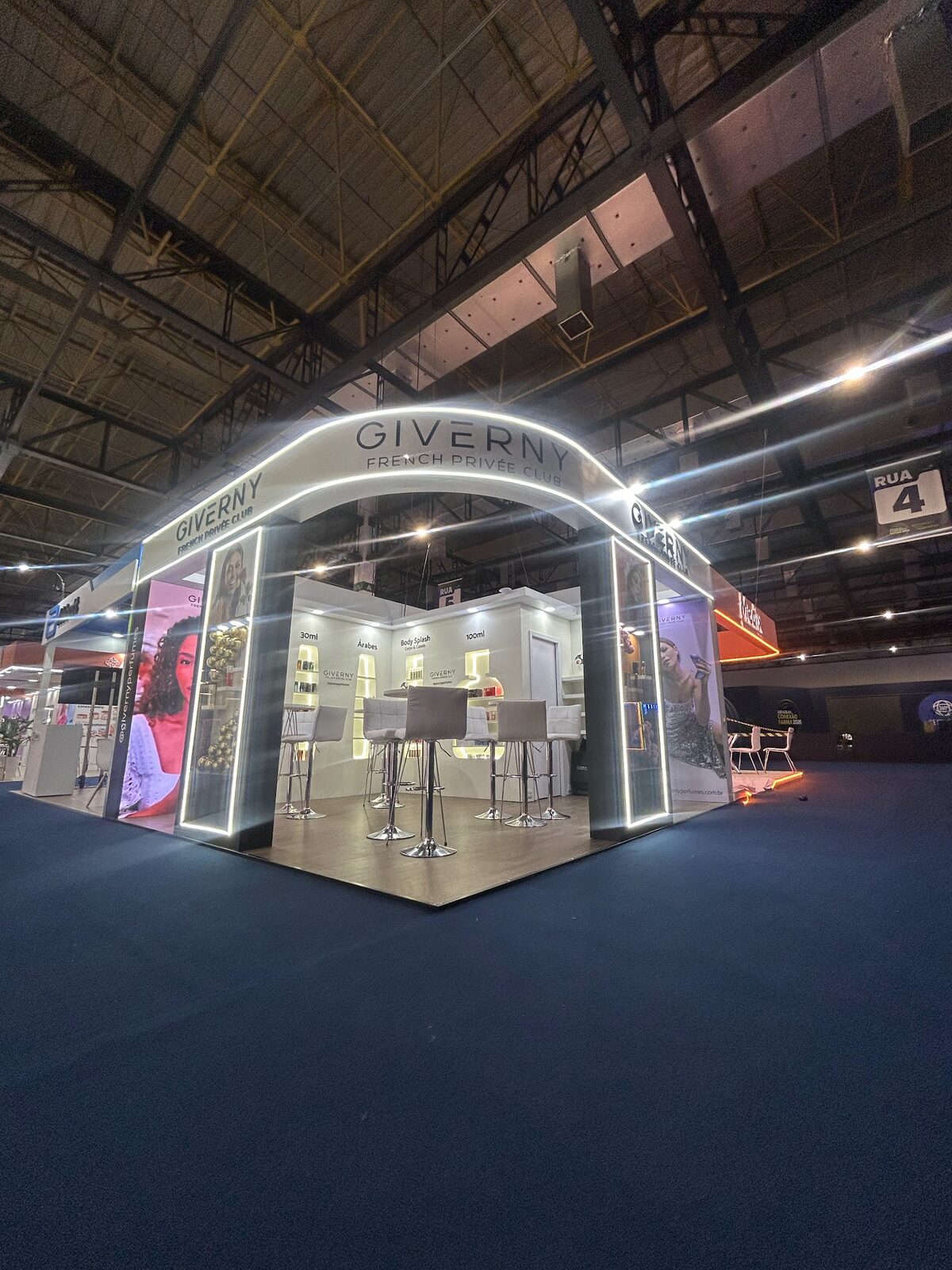 Stand de perfumaria com pórtico arqueado iluminado e prateleiras de produtos em pavilhão de feira