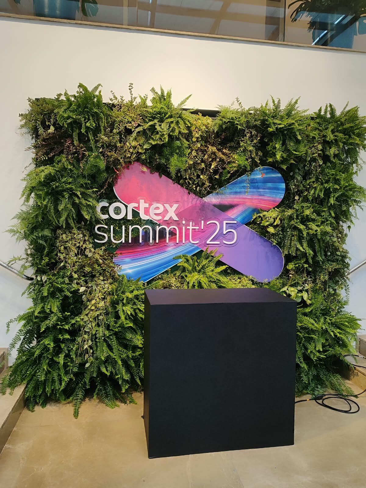 Parede de samambaias com marca em destaque, formato usado como backdrop cenográfico em evento corporativo