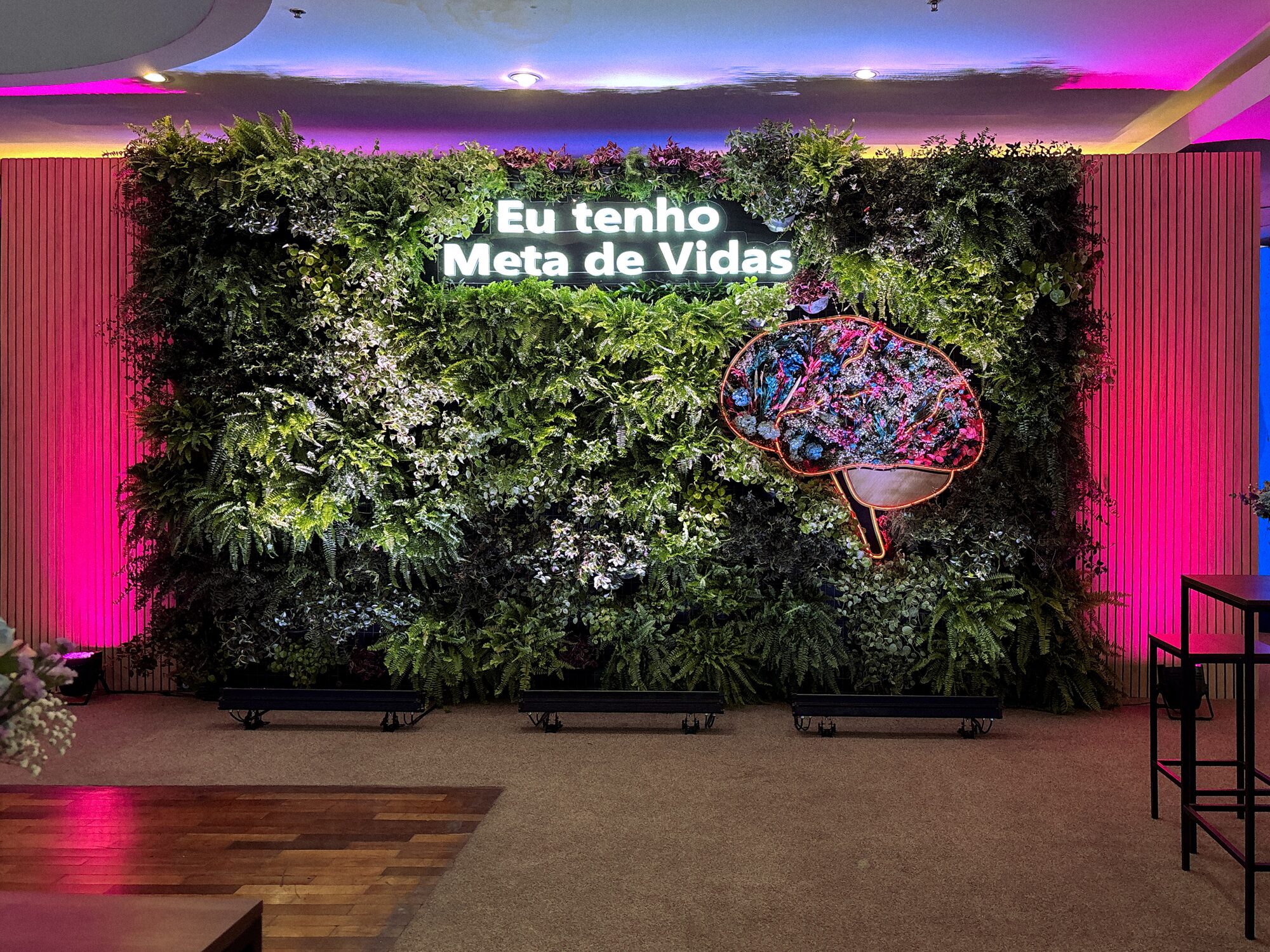 Backdrop com jardim vertical, letreiro neon 'Eu tenho Meta de Vidas' e escultura de cérebro em botânicos preservados