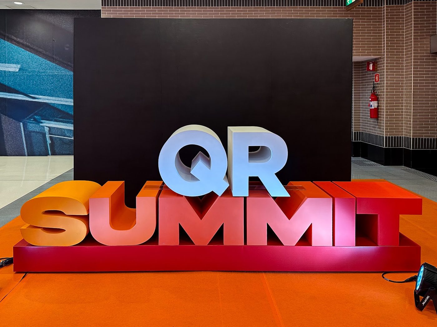 Escultura cenográfica 3D com letras QR Summit sobre tapete laranja em evento corporativo