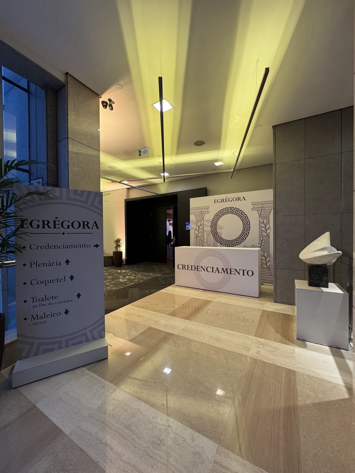 Área de credenciamento com balcão cenográfico, totem de sinalização e escultura em evento corporativo
