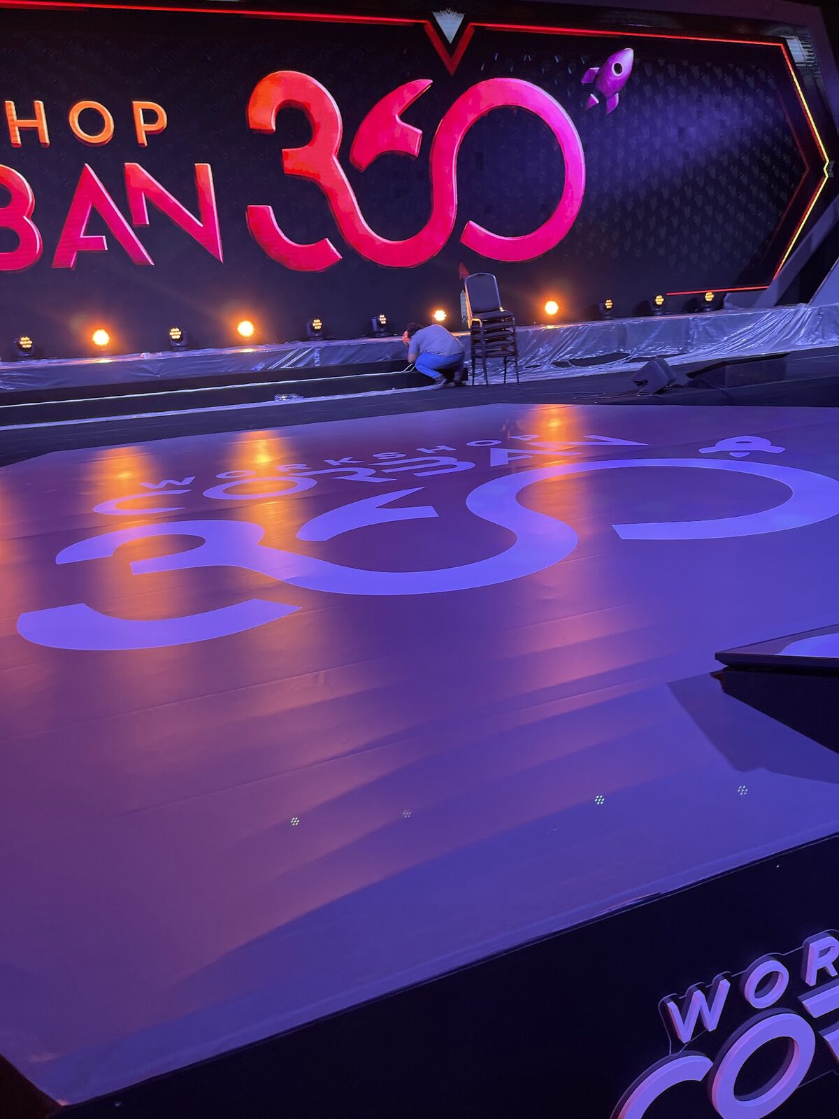 Palco cenográfico em fase de montagem com piso refletindo o logotipo do evento corporativo