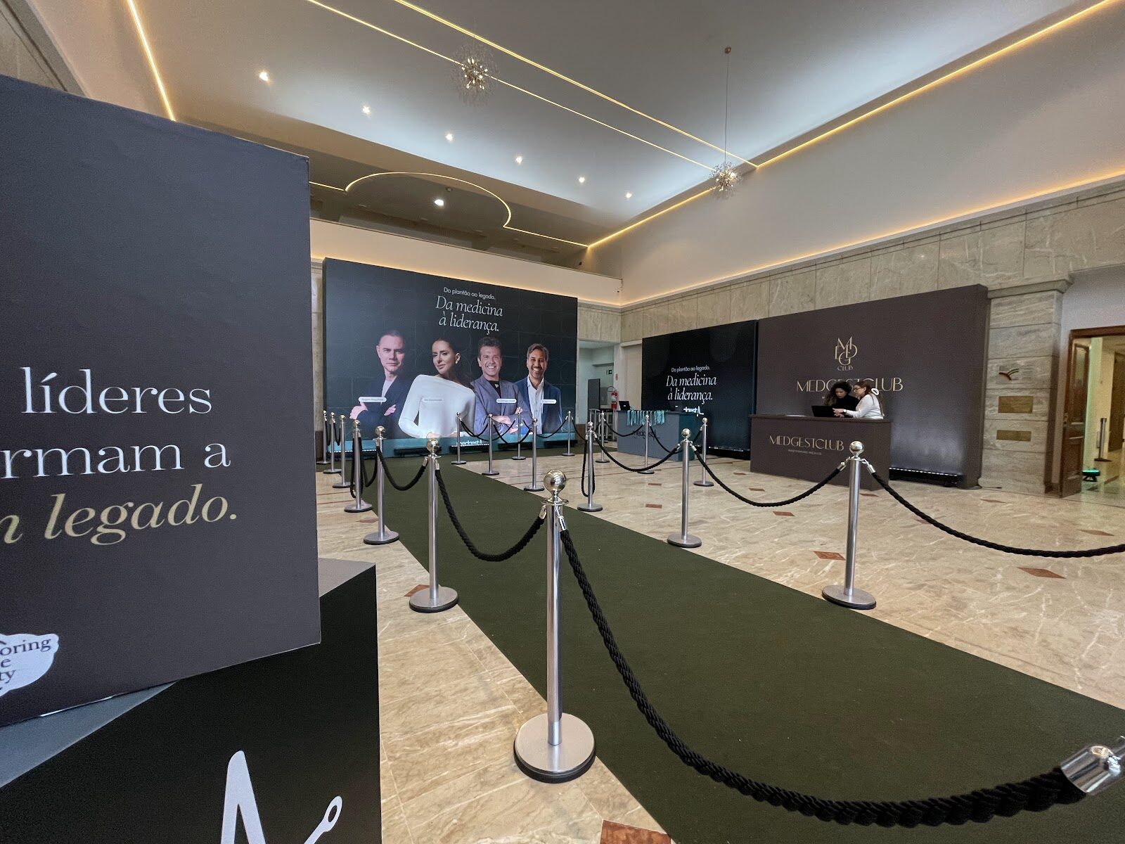 Lobby de evento corporativo com recepção, backdrop de marca, tapete e cordões de isolamento