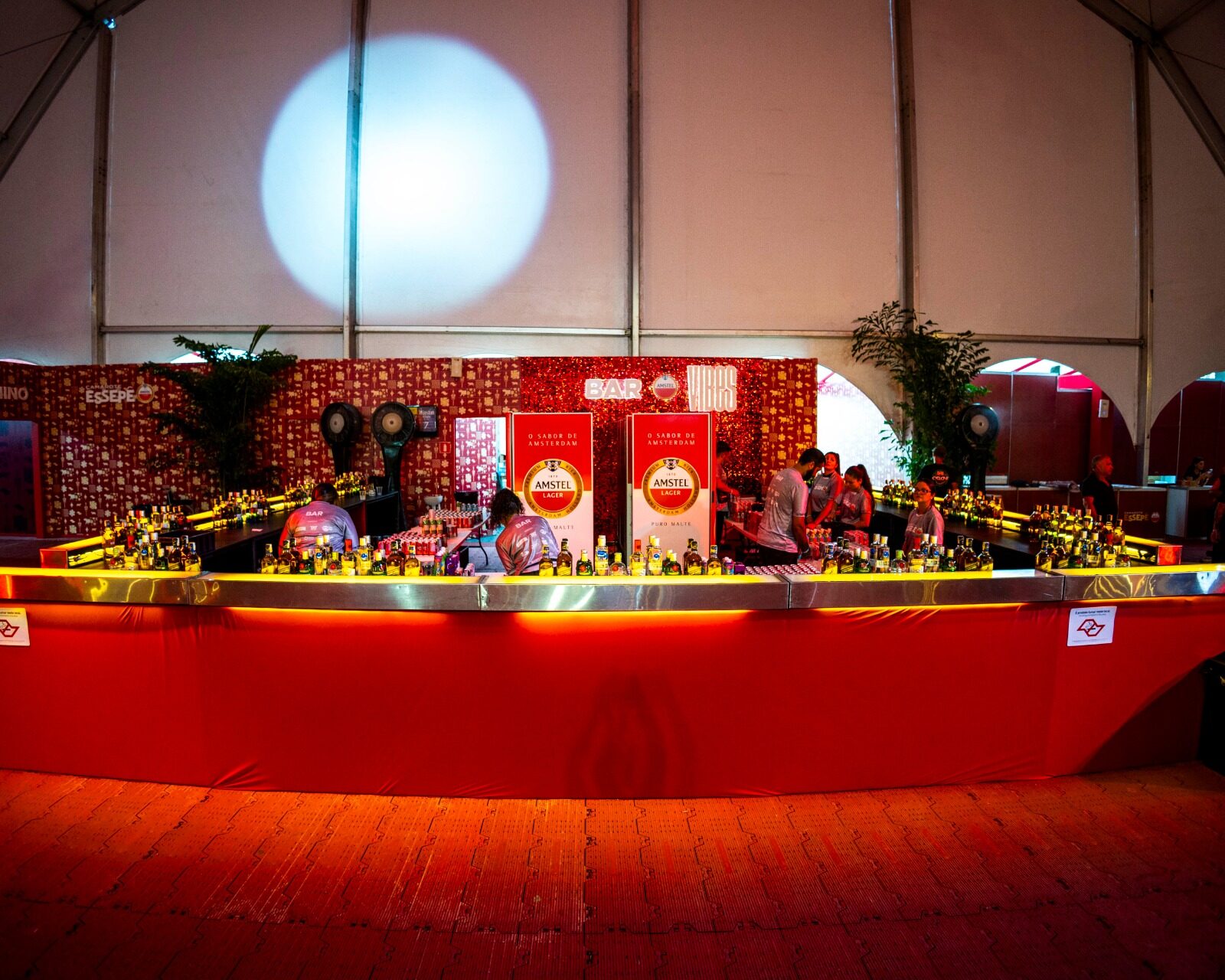 Bar circular vermelho iluminado em cenografia de evento, com bancada contínua de serviço para degustação de bebidas