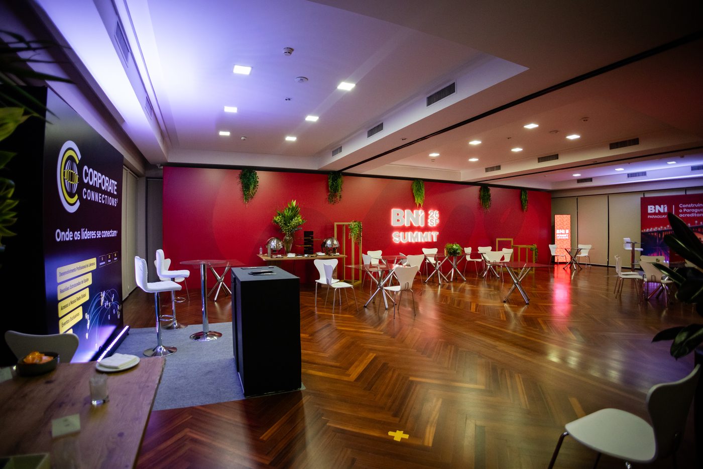 Lounge de networking do BNI Summit 26 SP com mesas altas, sinalização de patrocinadores e ambientação em vermelho e branco