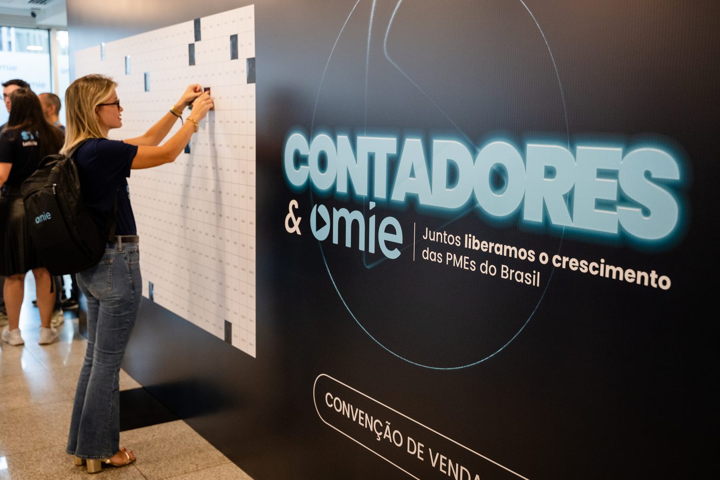 Participante interagindo com backdrop de agenda ao lado do painel Contadores & Omie na convenção