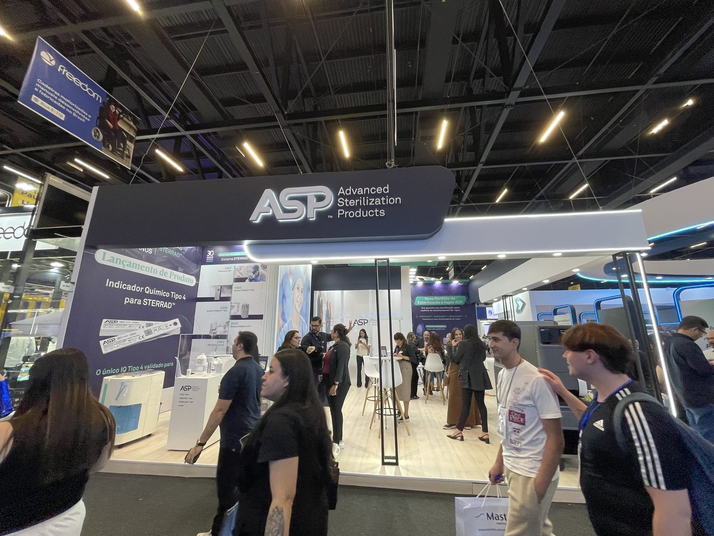 Estande de esquina da ASP na Feira Hospitalar, com testeira curva retroiluminada e piso amplo com mesas altas