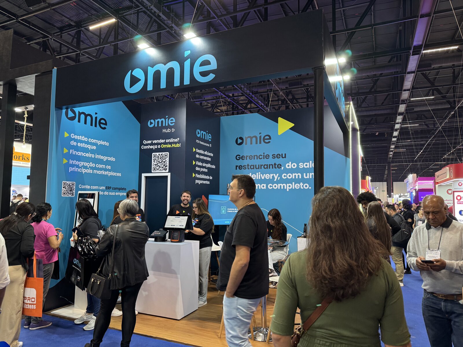 Área de convivência do estande da Omie na Fispal Food Service 2025, com mesas bistrô para demonstração do produto, montado pela Atom Cenografia