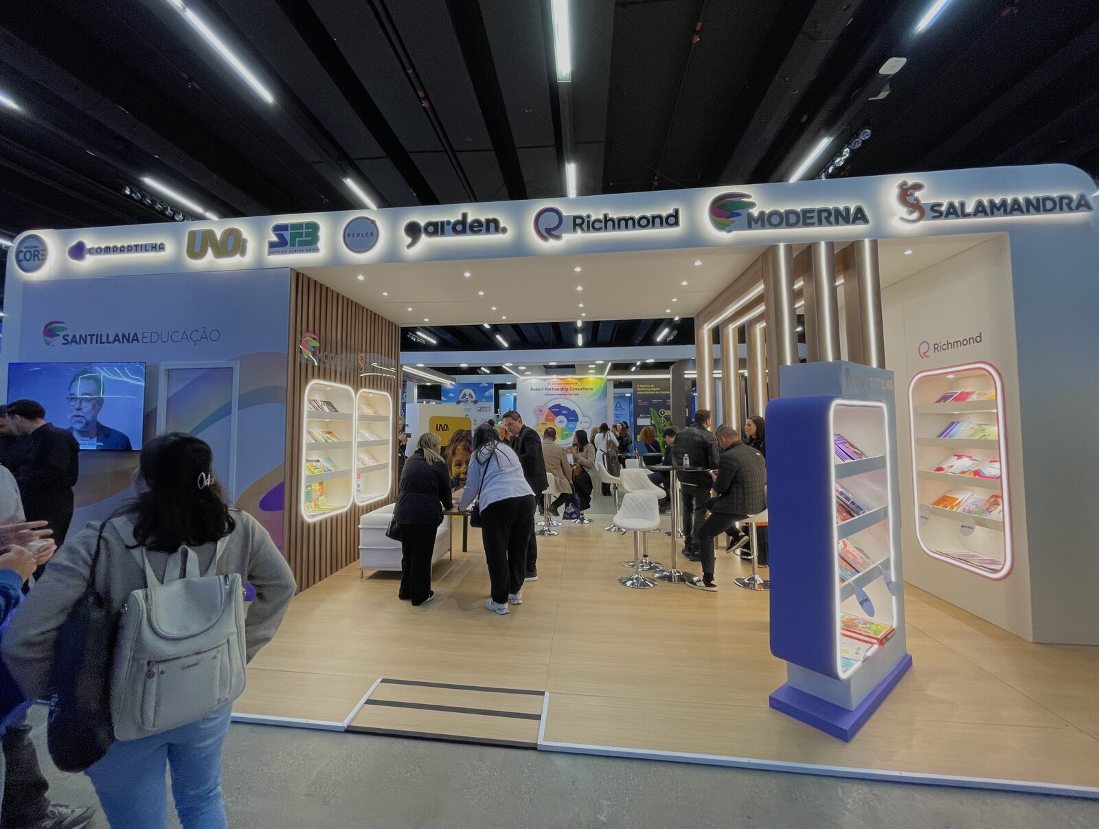 Stand ilha de 72 m² da Santillana Educação no 21º Crescer, com testeira reunindo os logos de dez marcas do grupo, montado pela Atom Cenografia